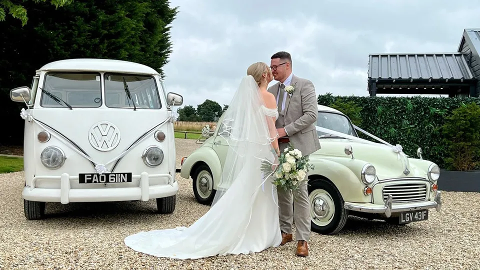 Classic VW splitscreen Campervan and a Morris Minor decorated with white ribbons at a local Keighley wedding venue with Bride and Groom standing in the middle of the vehicles. Bride wears a white dress holding a bridal bouquet and groom wears a cream suit