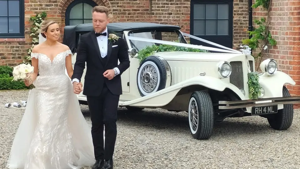 1930s vintage style convertible Beauford with black soft-top roof up in front of Wakefield wedding venue. Vehicle is decorated with ivory Ribbons and Bride and Groom are holding hands in front of the vehicle.