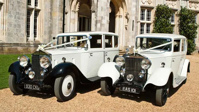 Pair of identical Vintage Rolls-Royce Limousine wedding cars in Black and Ivory decorated with traditional white wedding ribbons and bow across their bonnets. Vehicle is parked in