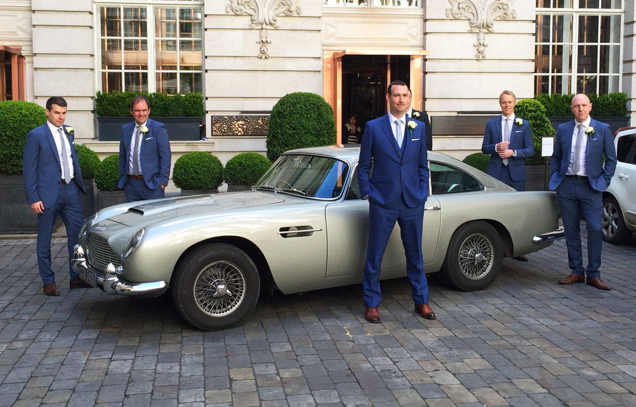 Classic Aston Martin DB5 in silver with groom and groomsmen around the vehicle posing for photos.