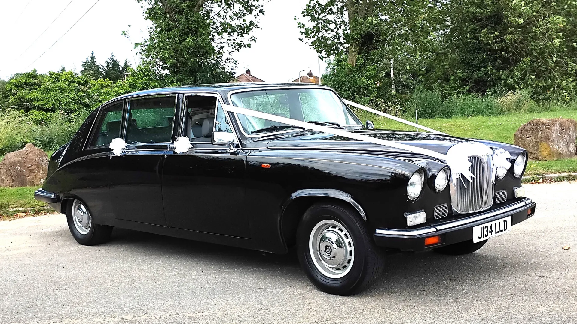 Right front view of Black Daimler Ds420 Limousine with white ribbons and white bows on door handles showing the twin headlights and Daimler Grill. Green forest background