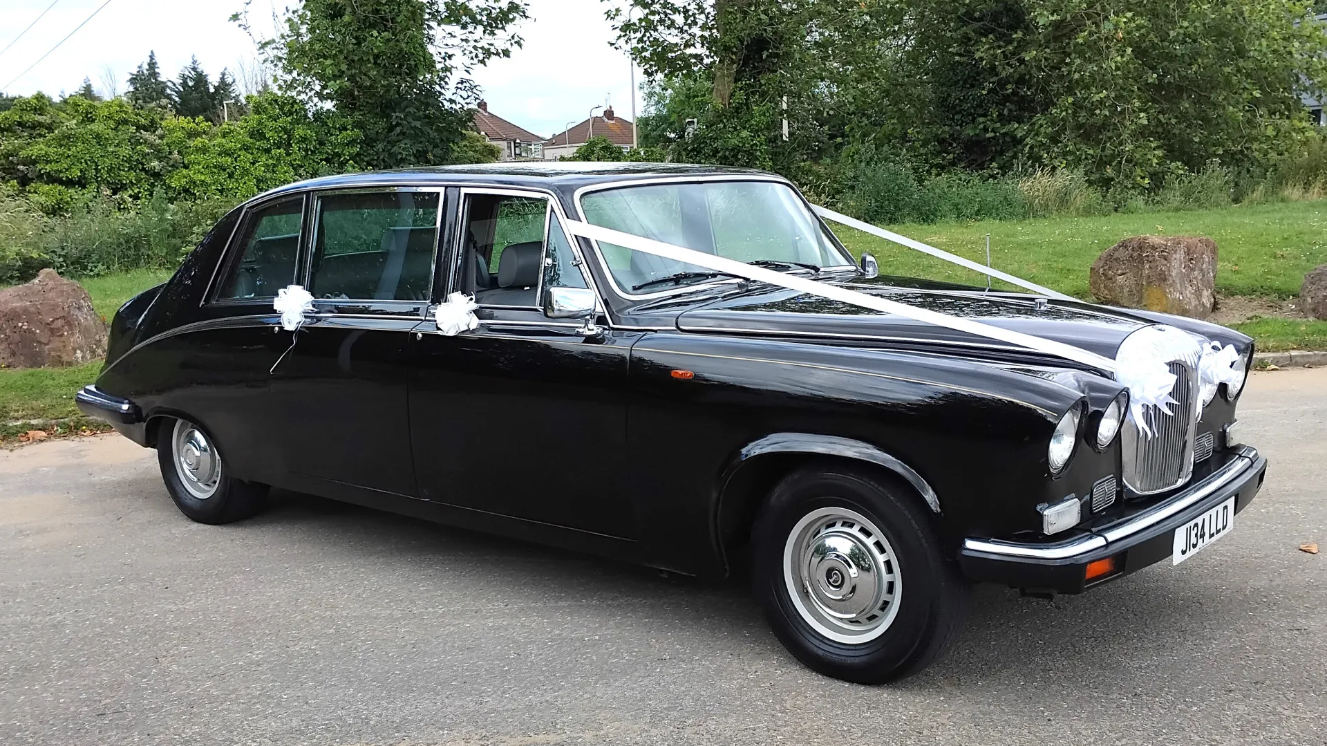 Right front view of Black Daimler Ds420 Limousine showing the twin headlights, Black Wheels and chrome wheel centre caps. Green forest background