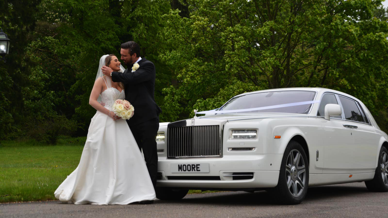 White Rolls-Royce Phantom decorated with white ribbons with Bride and Groom standing in front of the vehicle