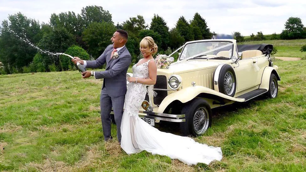 Ivory Beauford Convertible with its black soft top roof down in a local Leicester park. Spare wheel mounted on the left side of the vehicle. Bride and Groom are standing in front of the vehicle smiling. Groom has opened a bottle of Champagne.