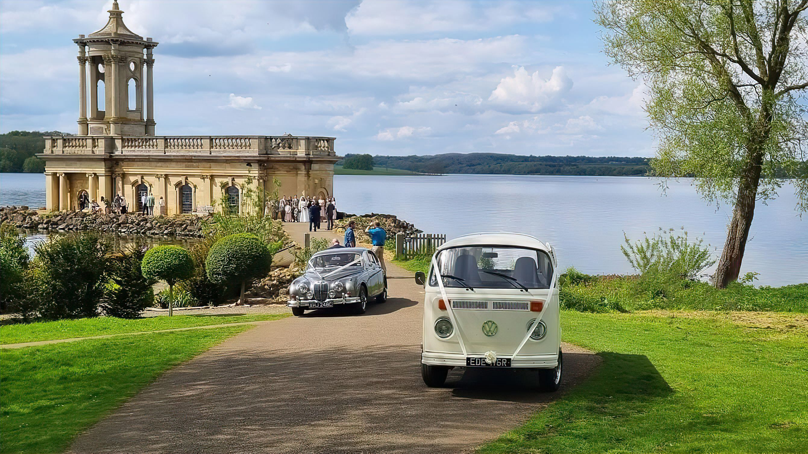 A classic VW campervan followed by a Jaguar Mk2 in front of Normanton Church near Leicestershire. Both vehicles are decorated with matching white ribbons. Wedding guests can be seen in the background.