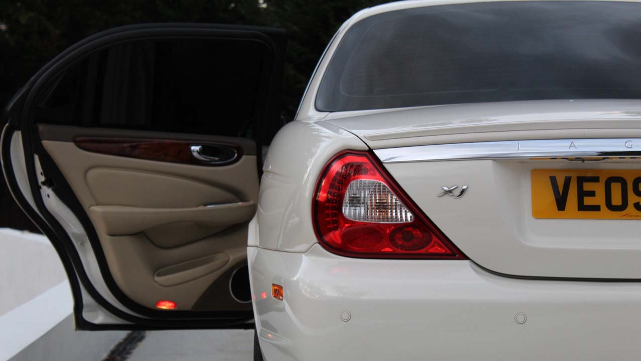 Rear view of White Jaguar XJ with rear left passenger door open showing cream leather interior on door card and wood panel on top of the door