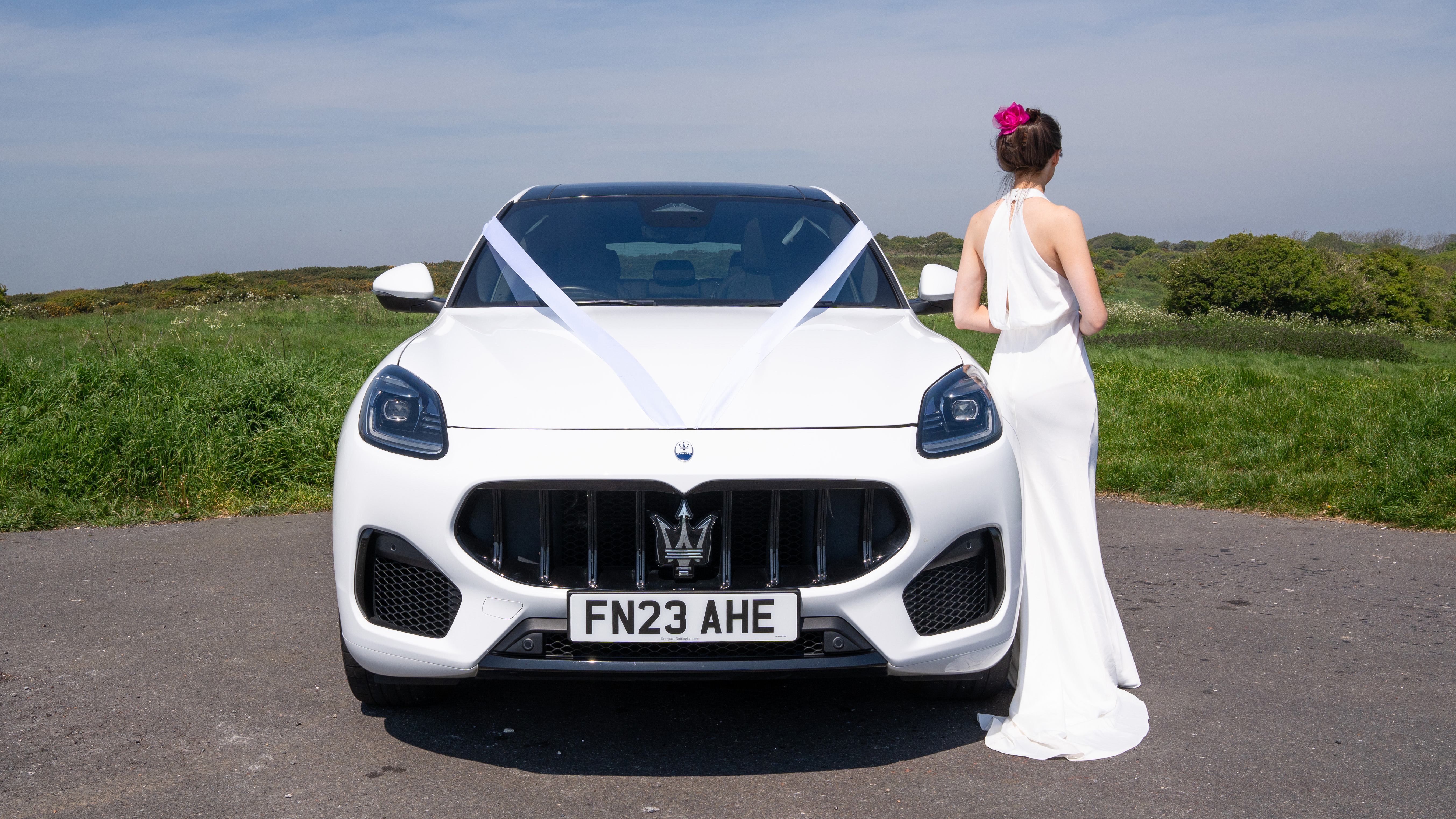 Full front view of White SUV Maserati Grecale with white ribbon, Maserati Trident Mascot in the middle of the front grill and Bride wearing a white dress leaning on the vehicle