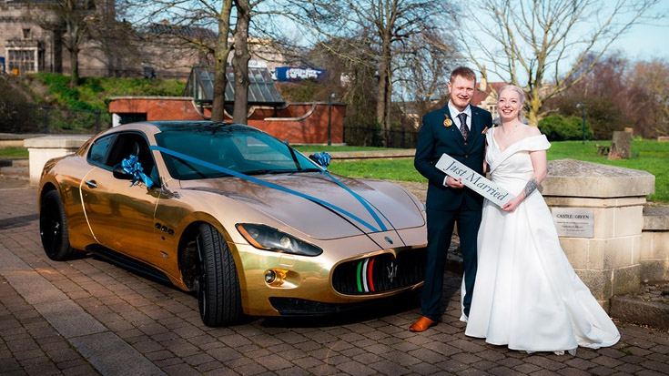 Gold Maserati with Dark Blue Ribbons. Bride and Groom are standing in front of the vehicle holding a "Just Married" sign