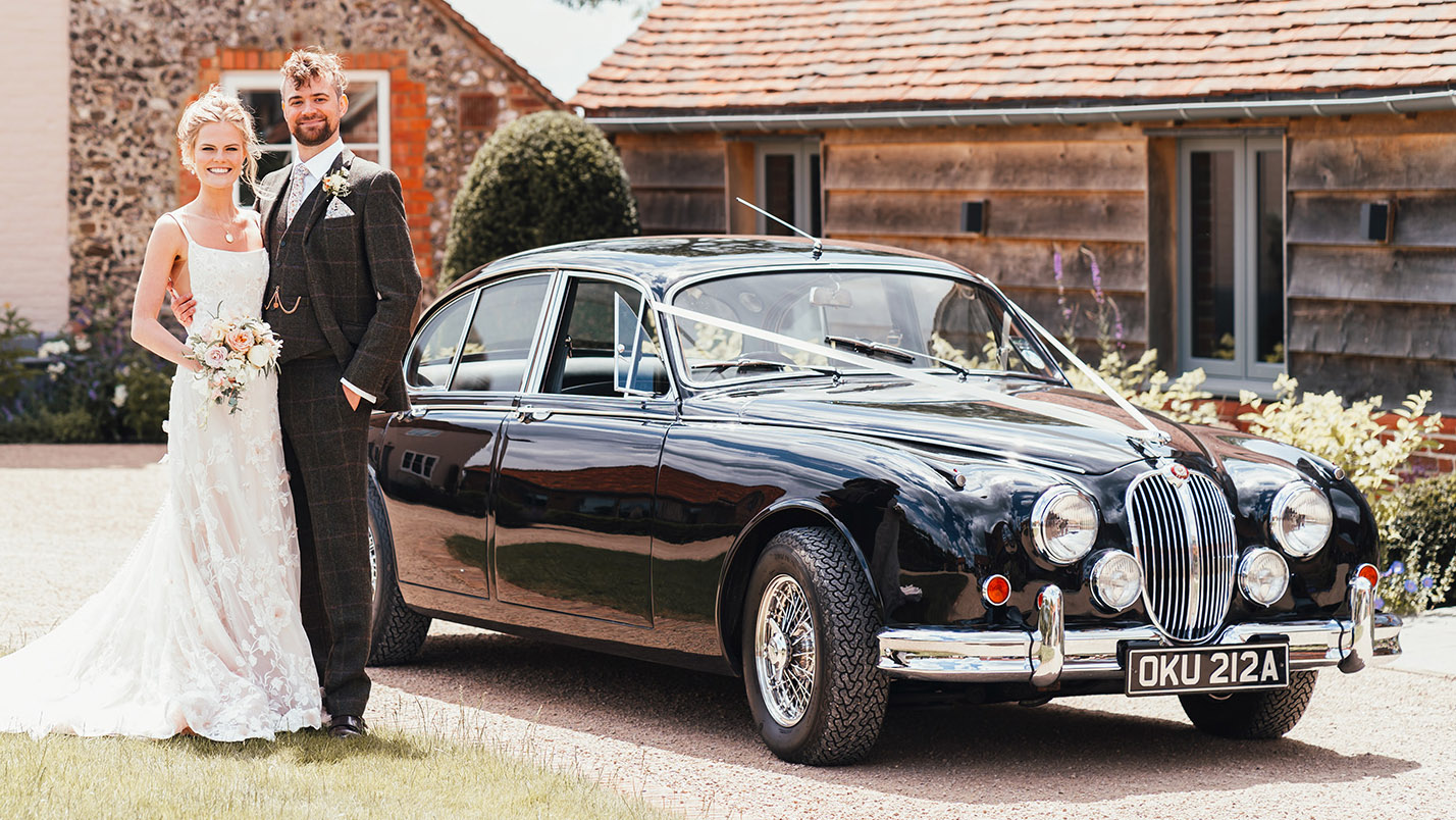 Classic Black RJaguar Mk2 decorated with White ribbons with Bride and Groom standing in front of the vehicle