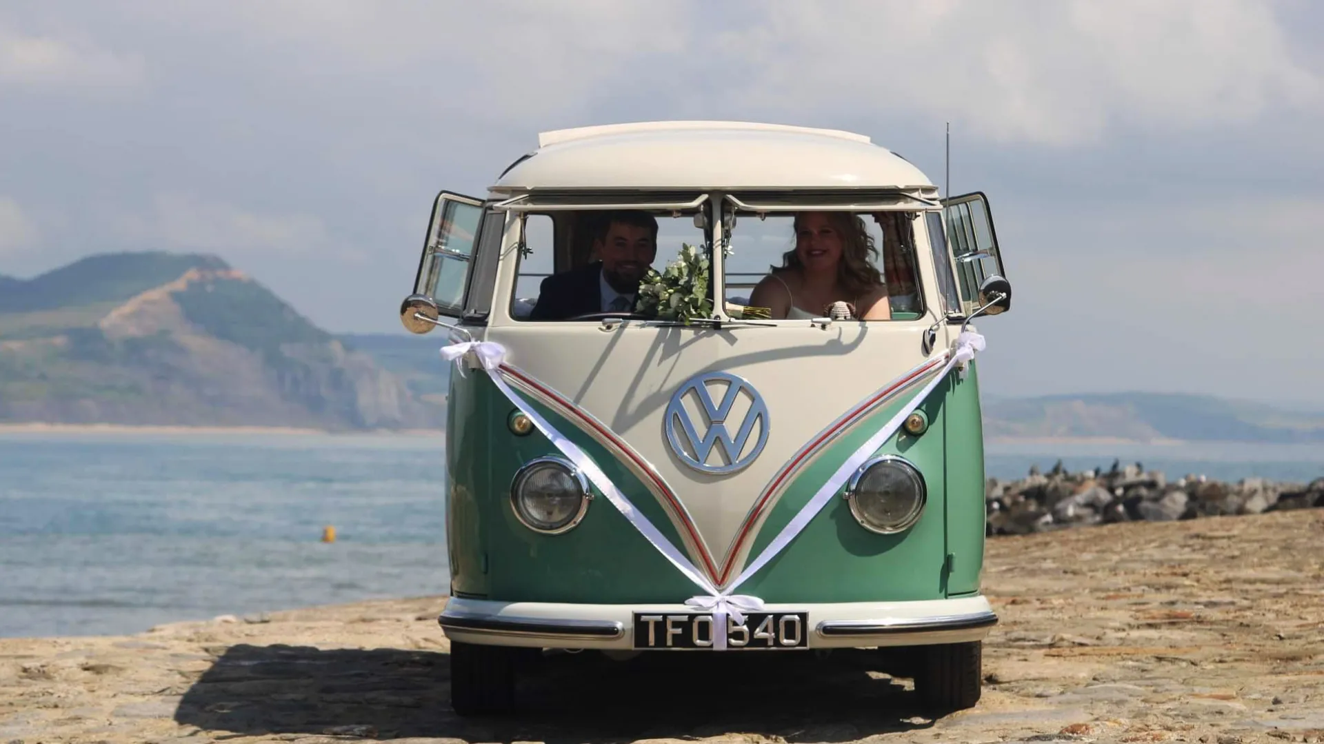 Full front view of classic campervan on Devon's seaside coast with Bride and Groom seating inside the vehicle looking through the open front window.