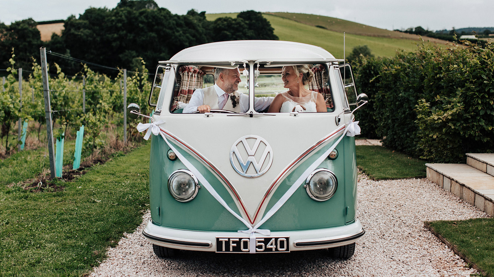 Full front view of classic campervan in Devon countryside with Bride and Groom seating inside the vehicle looking through the window.