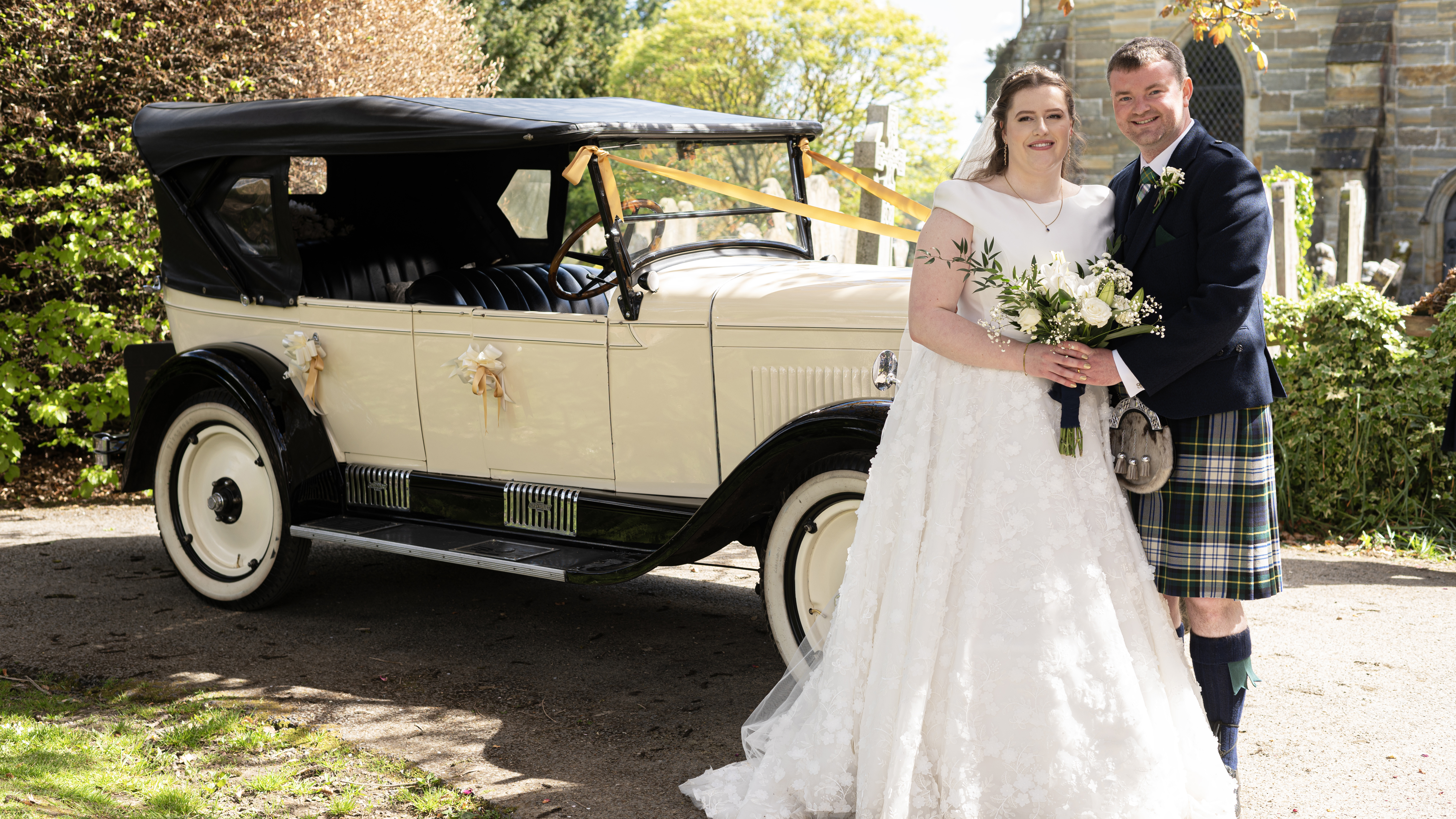 Bride and Groom posing for photos in front of a Vintage Chevrolet Convertible. Bride is wearing a white dress and holding flowers while the groom is wearing a Kilt