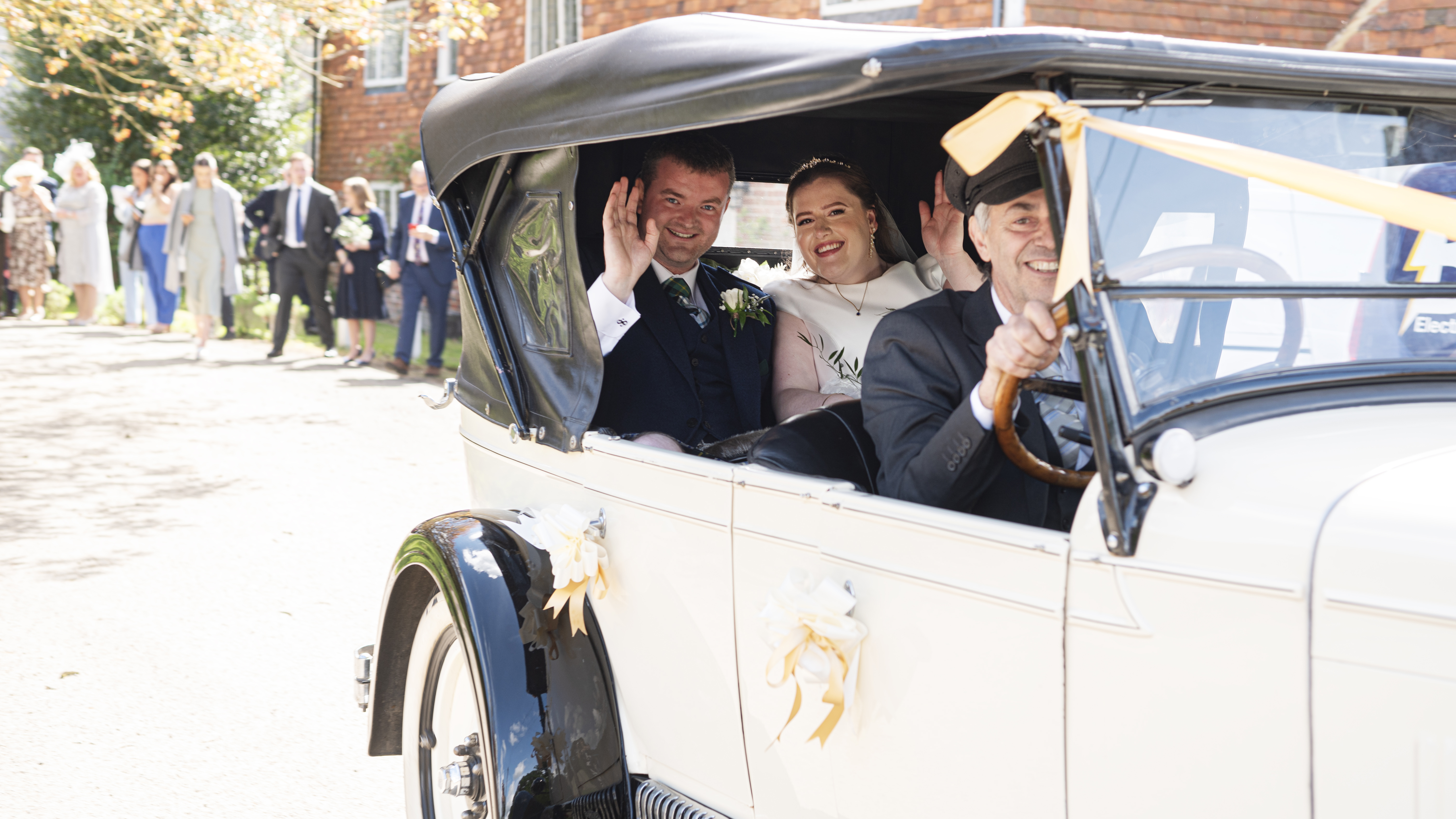 Bride and Groom seating in the back of the Chevrolet with smiling chauffeur driving. Side windows are down. Wedding guests can be seen in the background