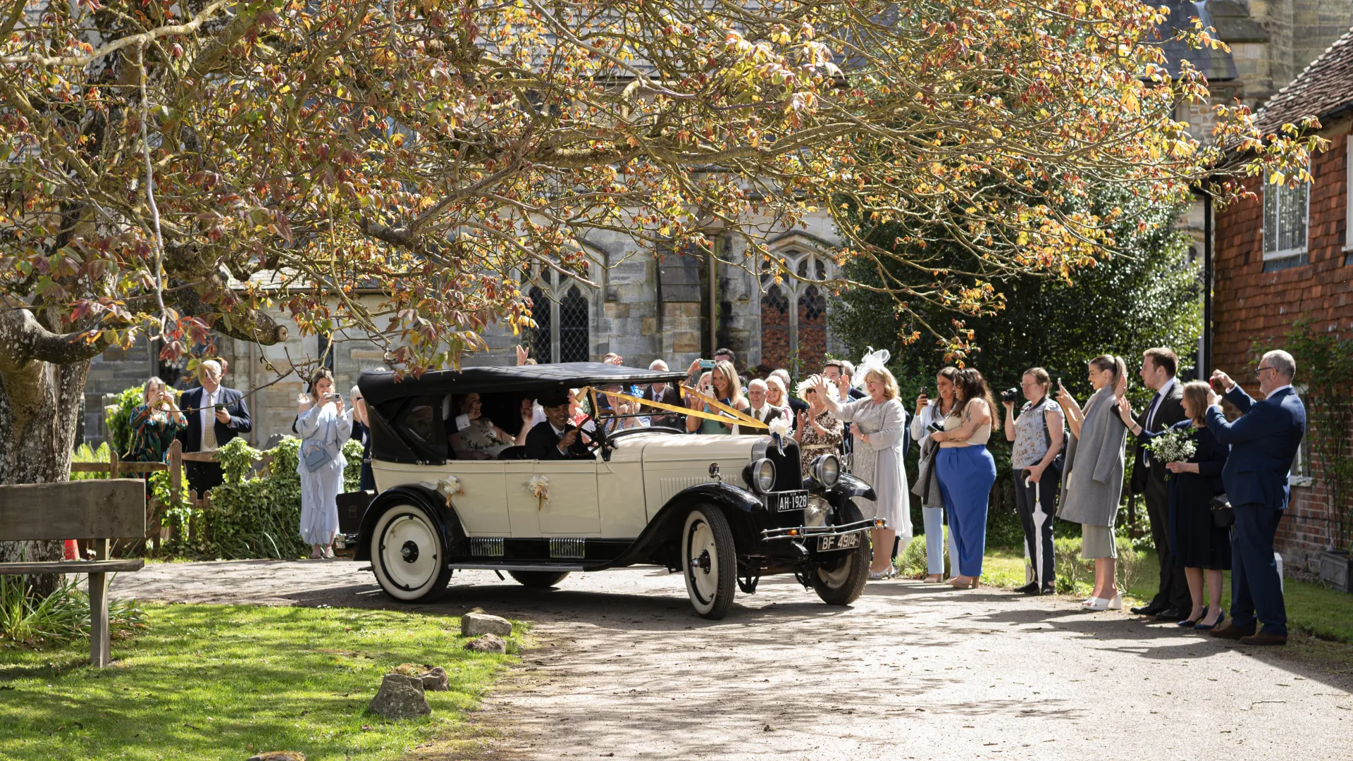 Vintage Chevrolet leaving the Church with Bride and Groom seating in the rear and wedding guests all around the vehicle wishing them farewell.