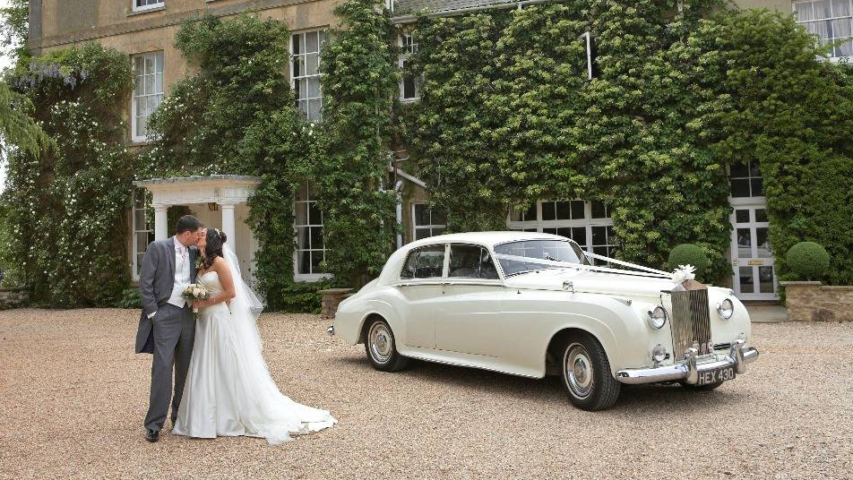 Classic Rolls-Royce decorated with ribbons in front of a wedidng venue with Bride and Groom holding each others next to the vehicle