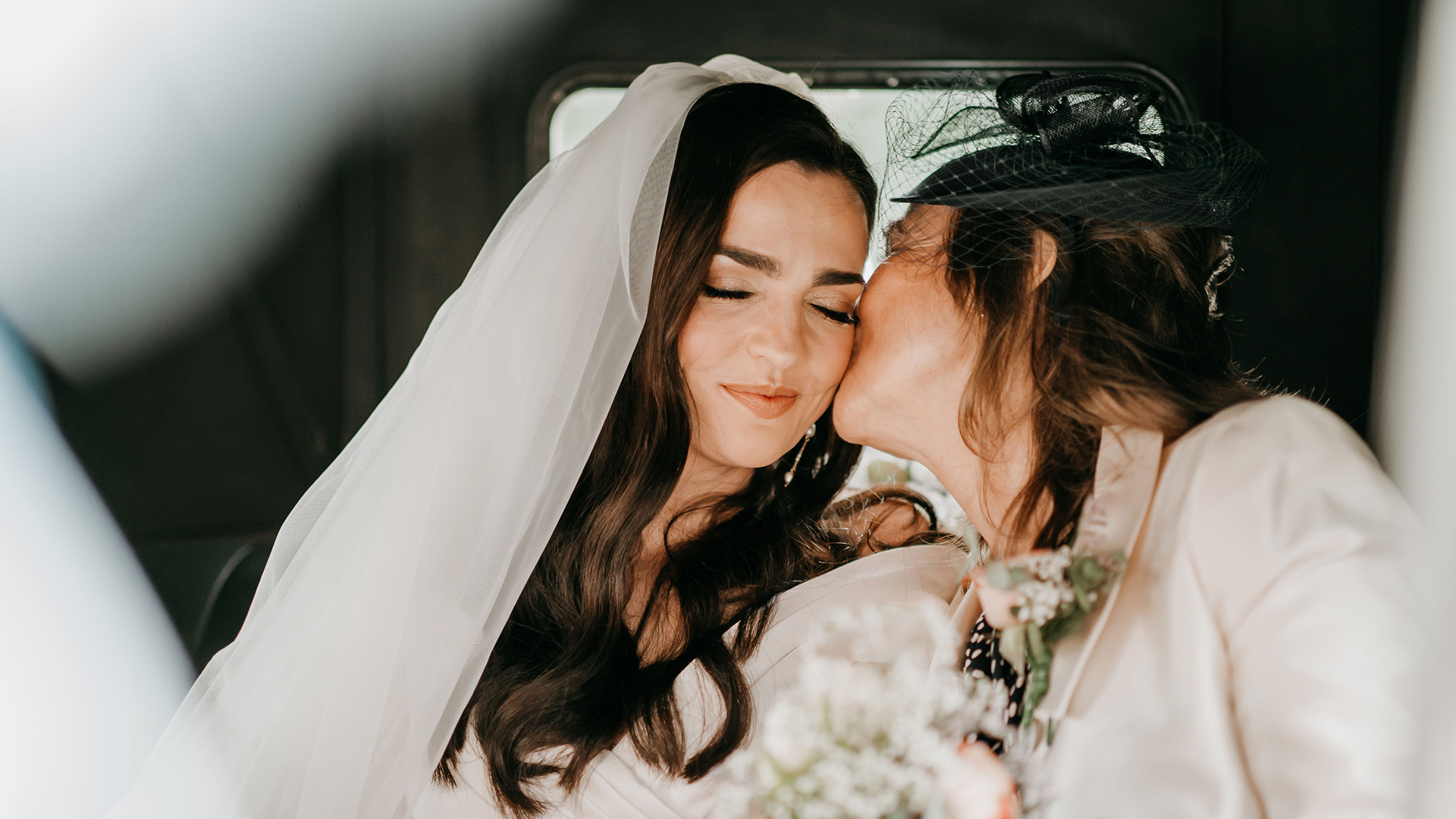 Bride and her Mother seating in the back of a vintage car. Mother is kissing Bride on her chick.