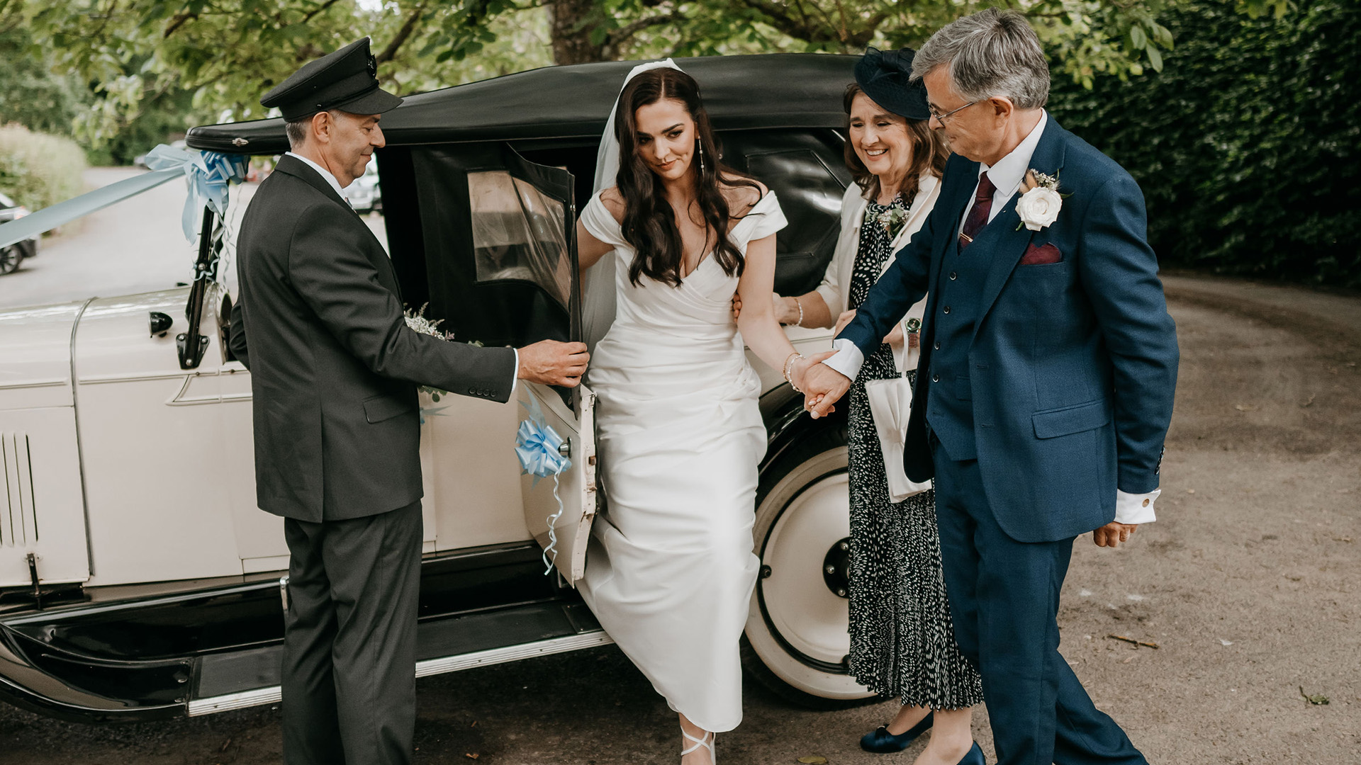 Fully suited chauffeur helping Bride with her white dress coming out of the vehicle. Brides parents are also on the photo