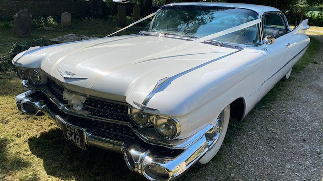 Front view of white cadillac with large chrome bumper. Car is decorated with white ribbons