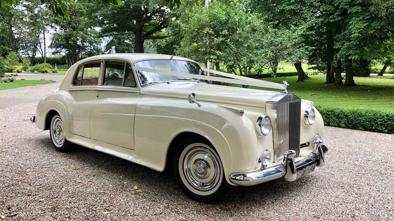 A Oxfordshire-based classic Rolls-Royce Silver Cloud in White dressed with traditional Ivory Ribbons across its front bonnet. Car has white wall tires and large chrome grill with s