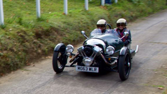 Morgan 3-wheeler being driven on road with 2 passengers wearing helmet