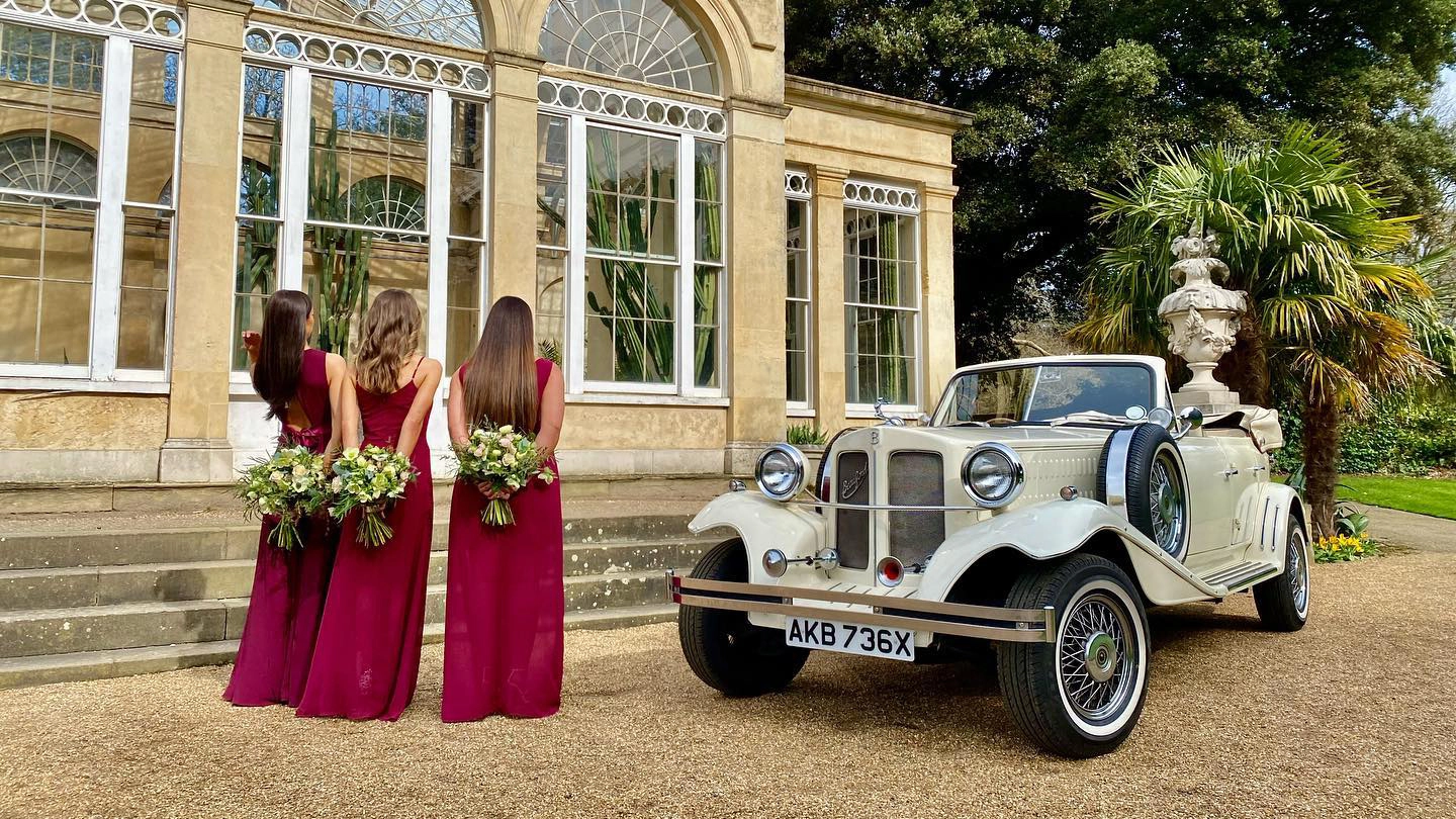 Vintage Beauford in Ivory with convertible soft top roof down and three Bridesmaids wearing a matching cherry-coloured dresses holding bouquet of flowers