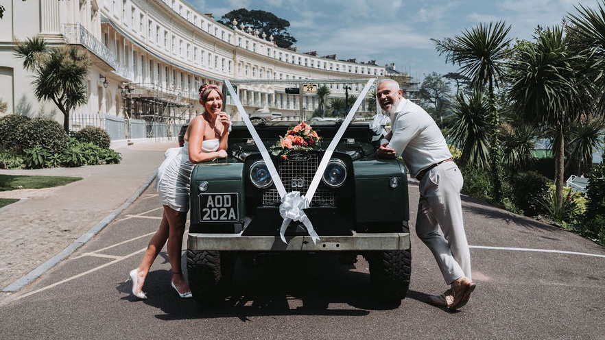 Bride and Groom standing on either side of the vehicle in front of Torquay wedding venue