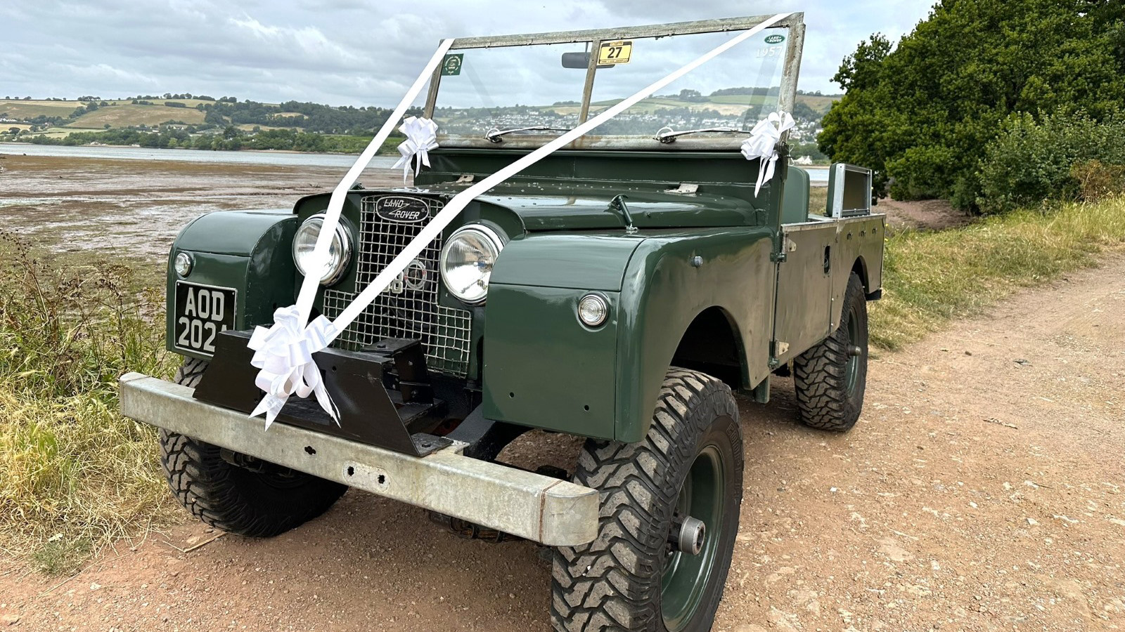 Front Left view of an Army Green Land Rover Series 1 decorated with White Ribbons and bows. Devon coastline can be seen in the background