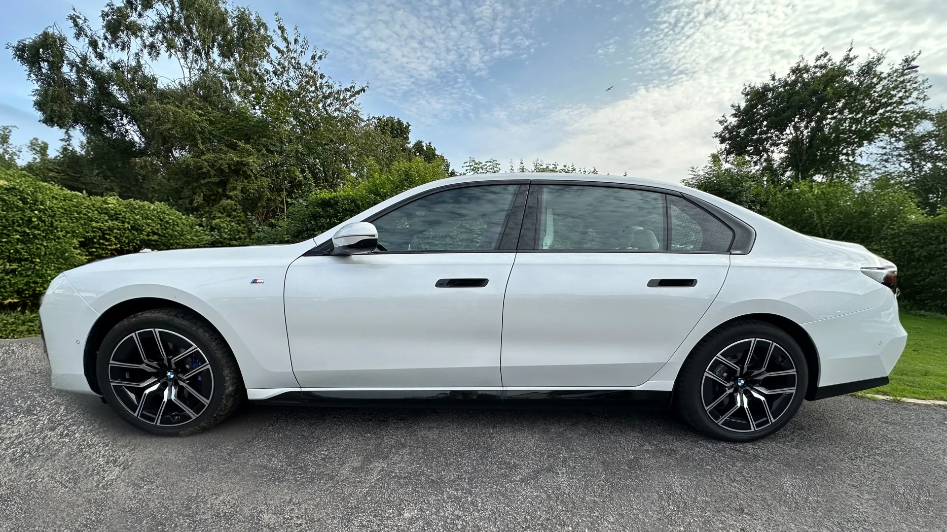 Left view of which Electric BMW i7 with Black and Silver Alloy Wheels, blue sky and green trees in the background