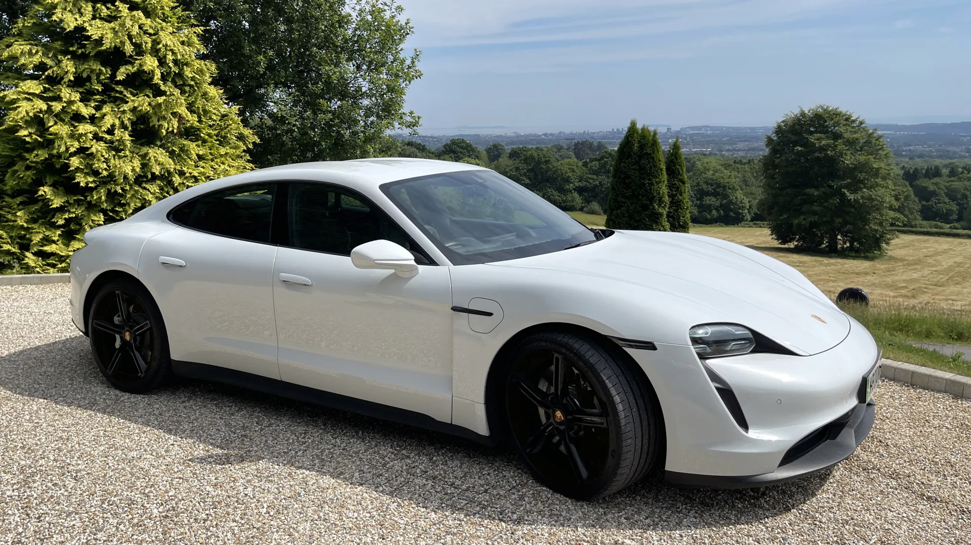 white Porsche Taycan Front side view parked in a driveway with blue sky and green trees in the background