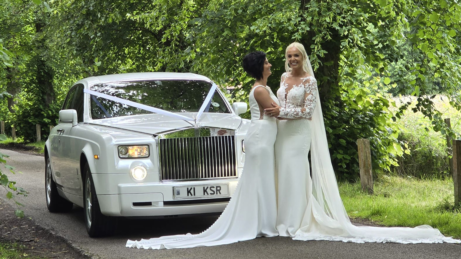 LGBT Gay Wedding with two brides in their wedding dress holding each others and smiling in front of a white rolls-royce phantom decorated with white ribbons