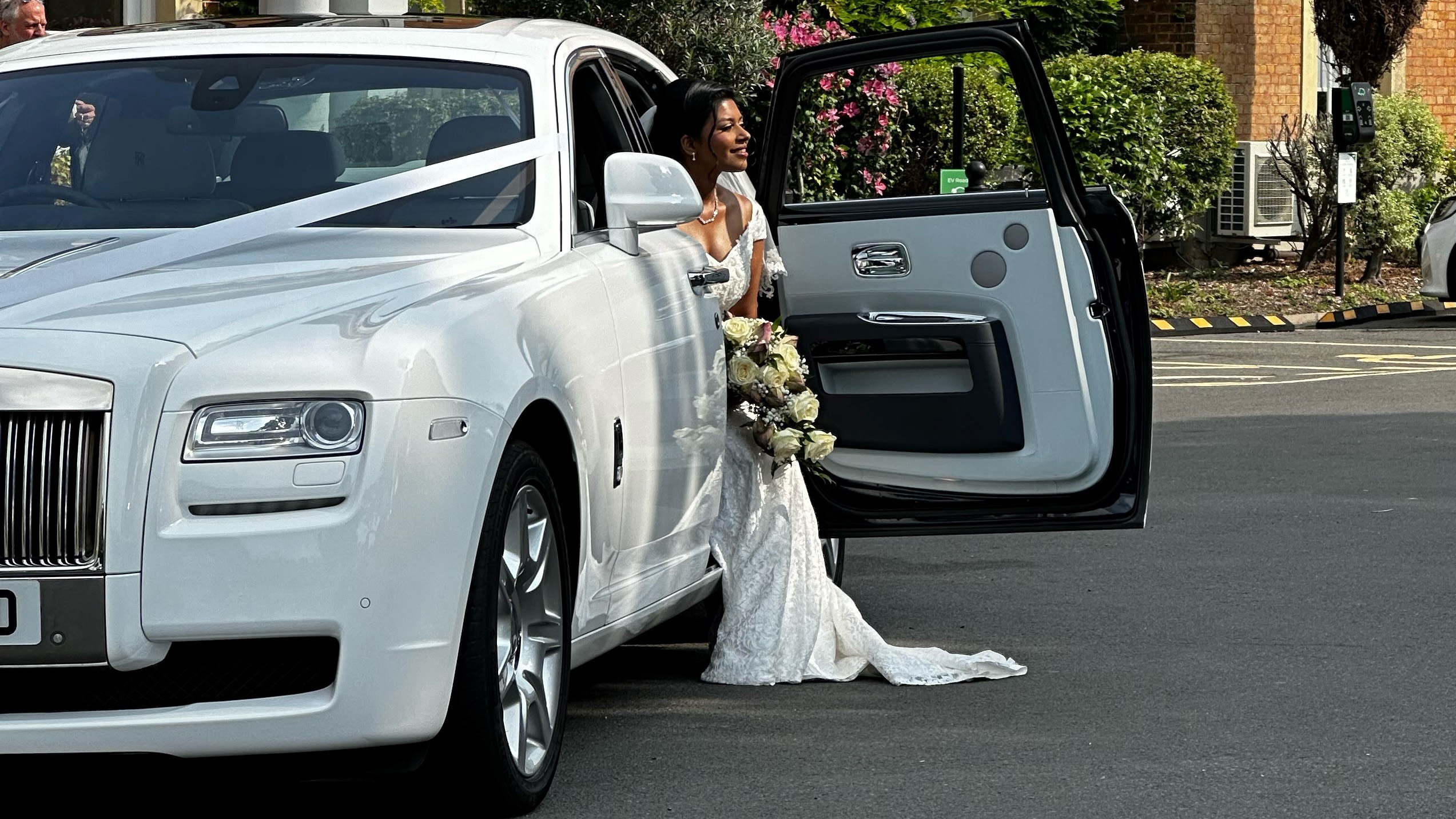 Bride wearing a white dress and holding a bouquet of flower exiting a White Rolls-Royce Ghost