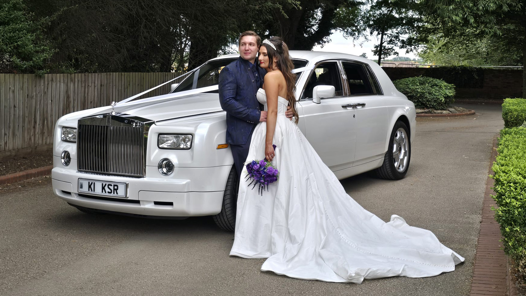 White Rolls-Royce phantom with ribbons. Bride and Groom standing in front of the vehicle holding each other.