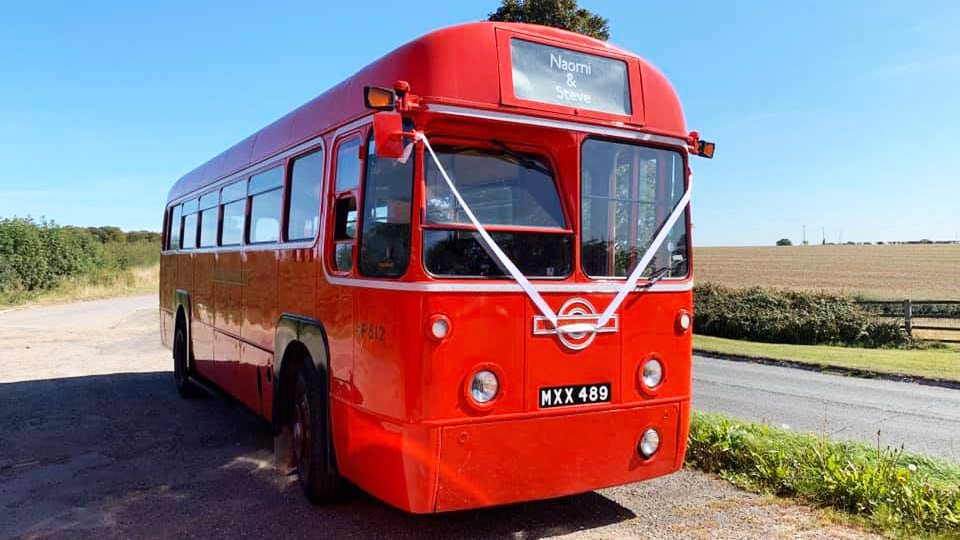 Right side front view of red single decker bus dressed with white wedding ribbons