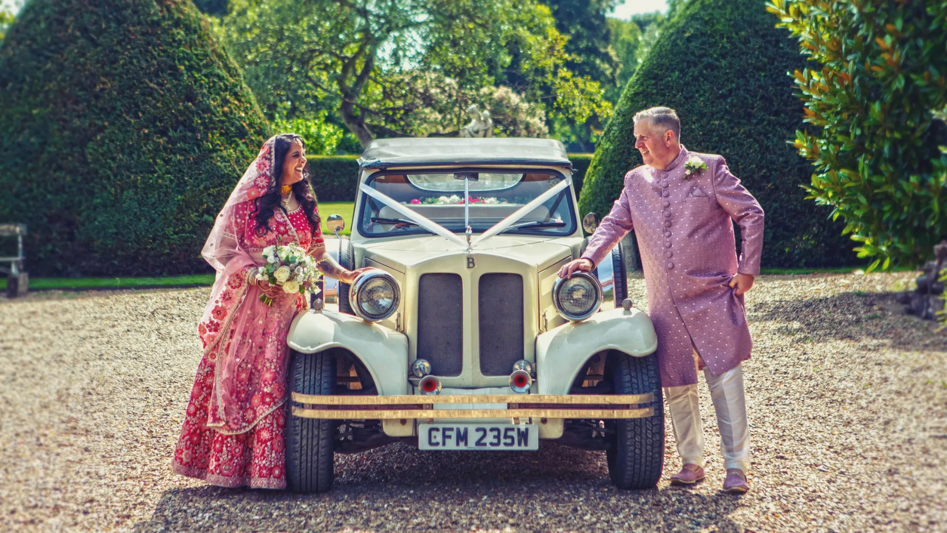 Asian couple in their traditional wedding outfit standing on either side of a Ivory vintage Beauford decorated with white ribbons