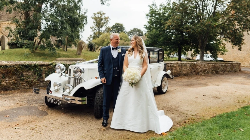 Bride and father standing in front of Vintage Beauford parked nest to the church. Bride is holding a large white bouquet of flower and her dad is wearing a navy blue suit with bow tie.