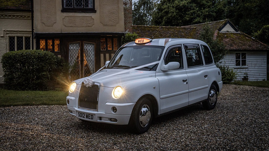 white taxi cab at Dusk with headlights switched on and orange sign on roof lit up showing "WED" wording