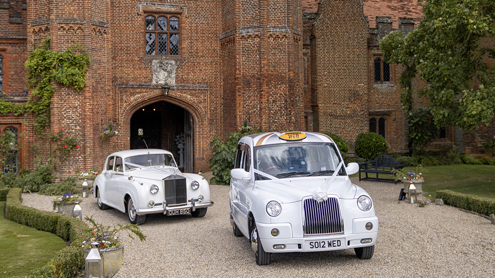 Two white wedding cars leaving the wedding venue in Essex. First car is a White taxi cab followed by a Classic Rolls-Royce both with matching white ribbons