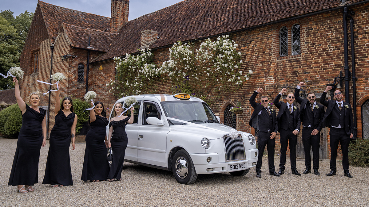 White taxi cab surrounded by Bridesmaids and Groomsmen wearing black outfits and cheering.