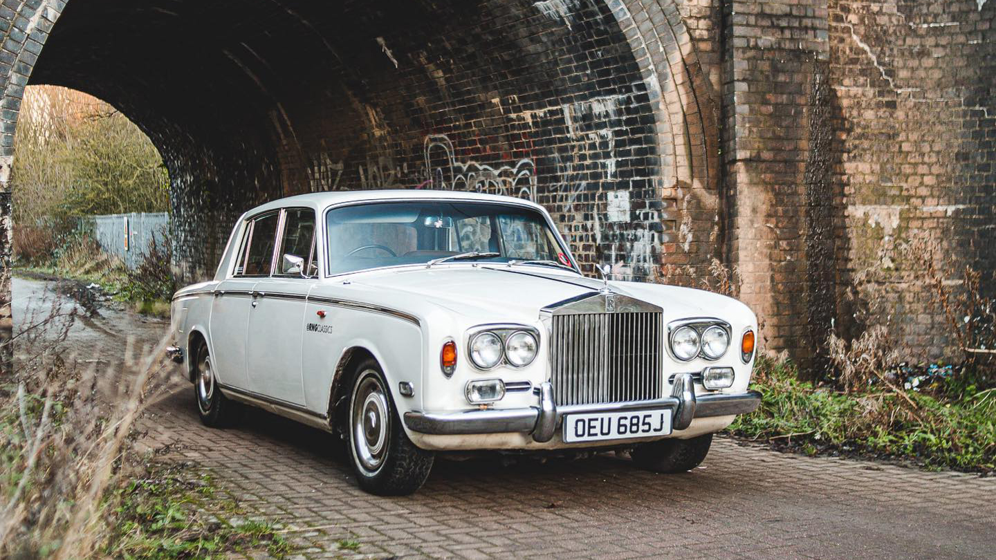 White Rolls-Royce Silver Shadow with twin headlights passing under a old brick bridge