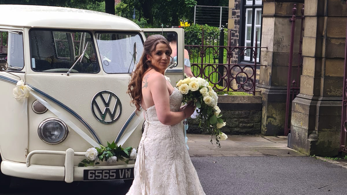 Bride holding a large bouquet of white flowers standing in front of classic campervan decorqated with white ribbons and bows