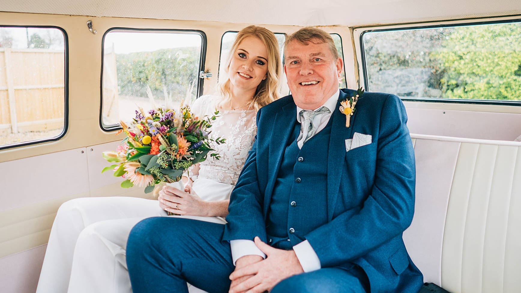 Bride and her father seated inside a vw campervan. Bride is holding a large colourful bouquet of flowers and her dad wearing a Turquoise blue suit