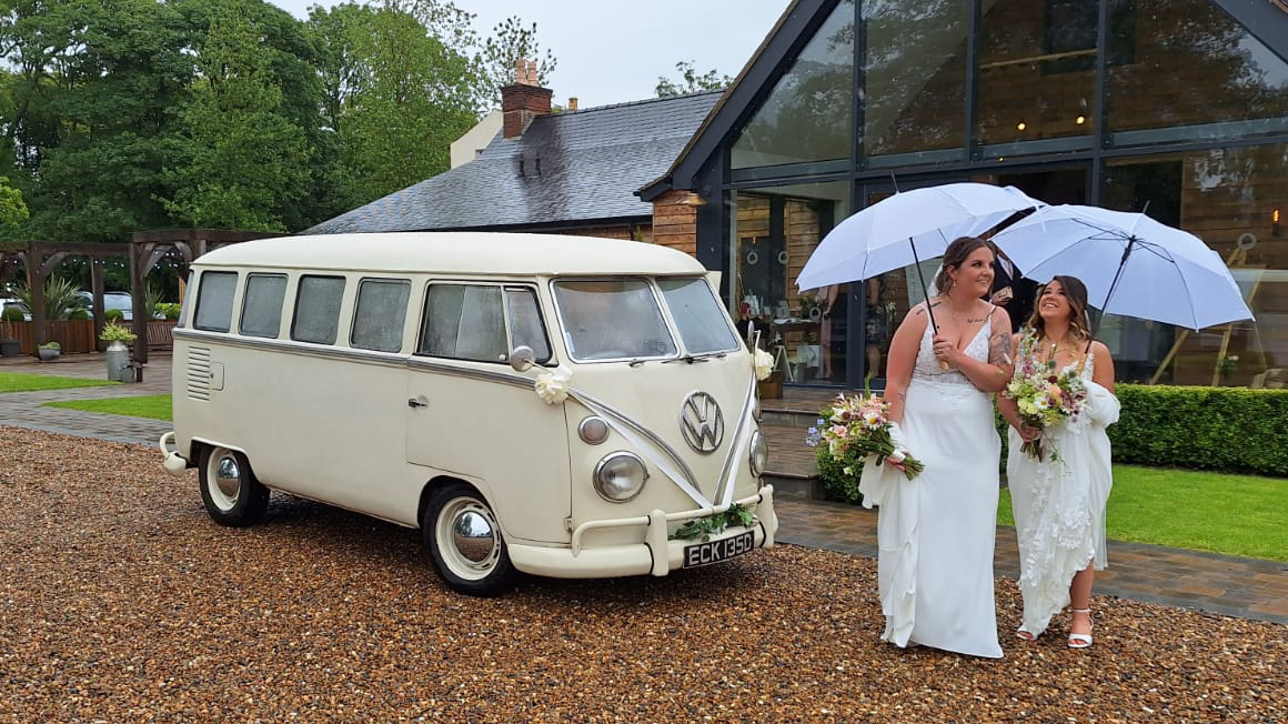 Two brides standing in front of classic volksagen campervan. Both brides are wearing white dresses and holding a large umbrella