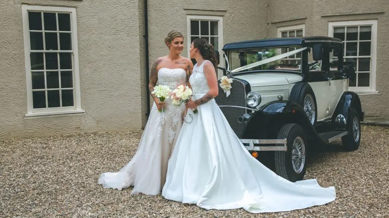 Two Brides smiling at each others in front of a Vintage Badsworth Landaulette with white ribbons