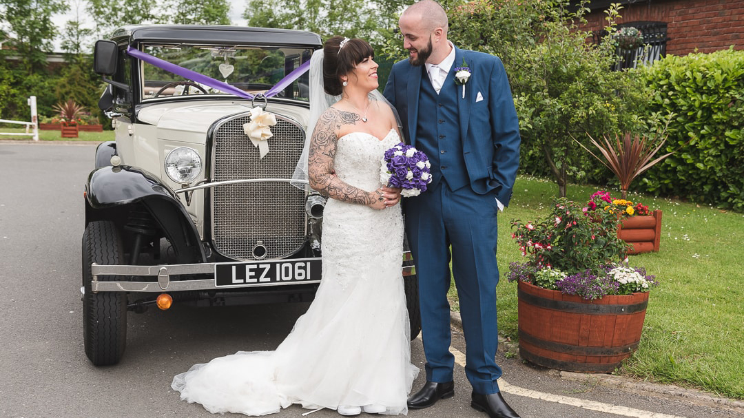 Bride and Groom standing in front of a vintage Badsworth decorated with purple ribbons