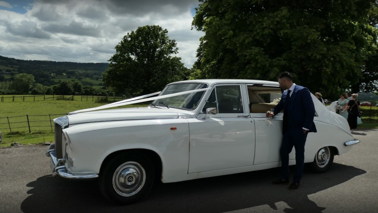 Classic Daimler Limousine in white on wedding duties with groom wearing a blue suit leaning on the car