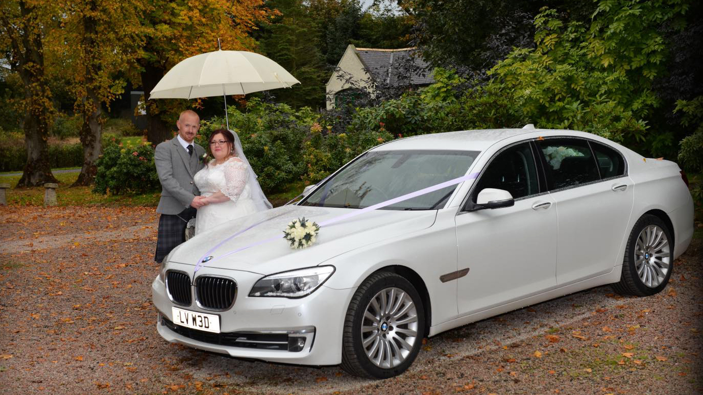 Bride and Groom under a white umbrella standing by a White BMW. autumn colours in the background