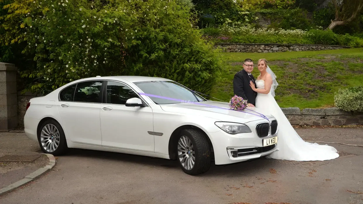 Bride wearing a long white wedding dress with her groom posing for photos next to a modern White BMW car