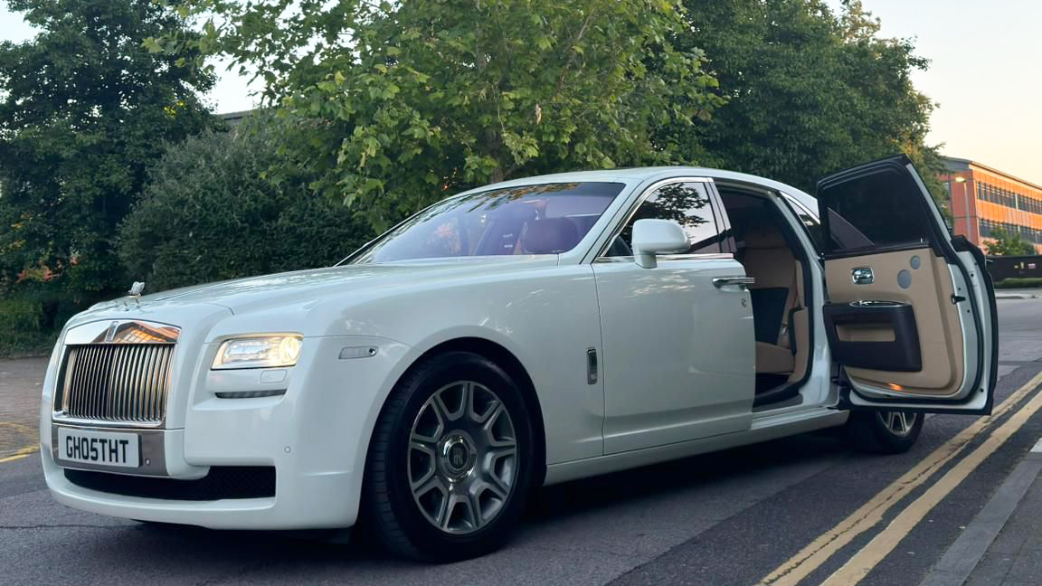 white Rolls-Royce Ghost with large chrome alloy wheels, forward opening rear door open showing leatrher interior and headlights switched on