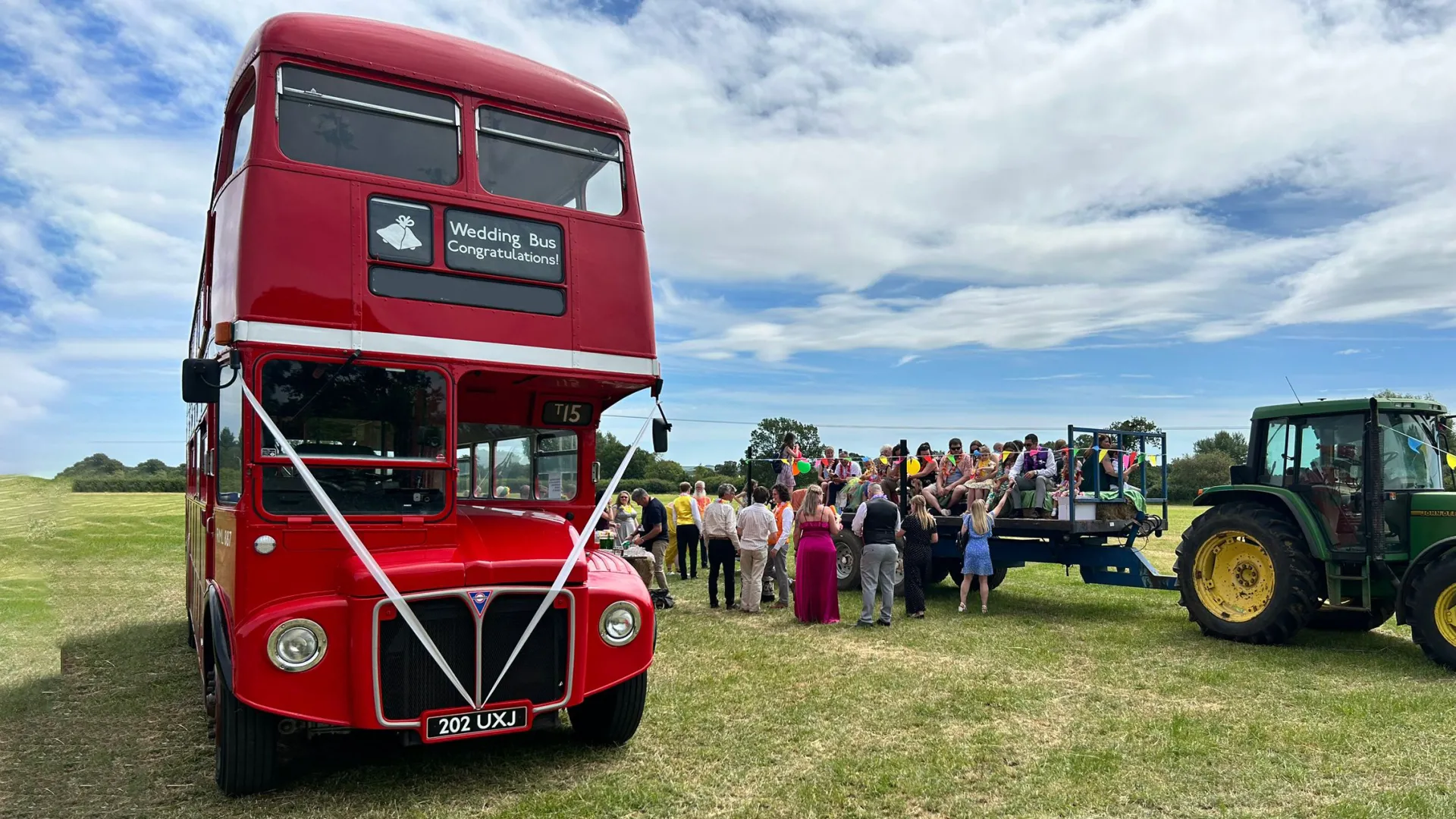 Red Old Routemaster Bus with white ribbons accross the front parked in a field next to a Green Tractor. Wedding Guests are seen in the background