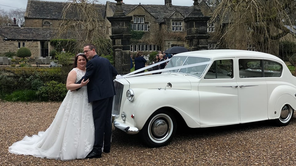 Bride and Groom holding each others in front of classic White Austin Princess wedding car.